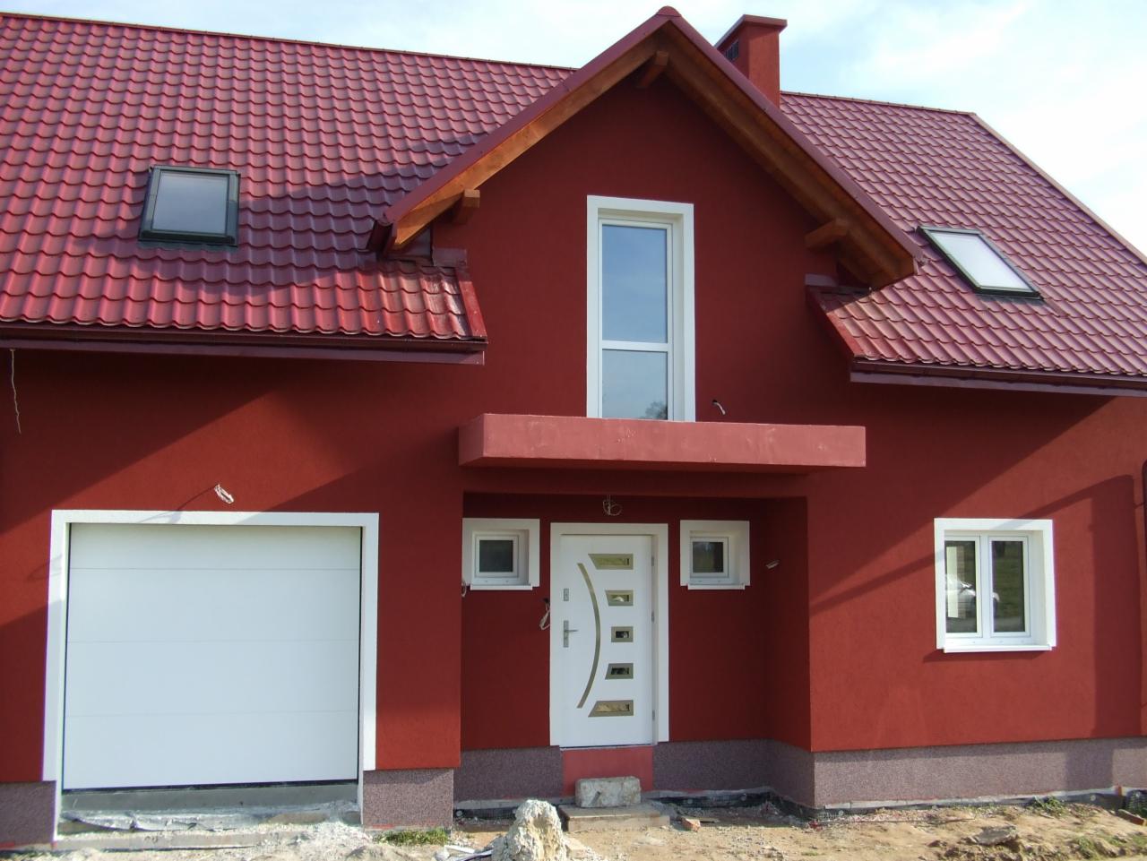 Czerwony dom jednorodzinny z białymi oknami i drzwiami garażowymi. Dach z czerwonej dachówki, okna dachowe. Widok z przodu, jasne niebo.