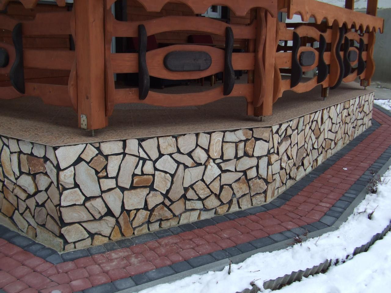 Wykończenie podstawy altany kamieniem elewacyjnym i kostką brukową w kolorach czerwieni i szarości. Zimowy krajobraz z elementami śniegu. Dekoracyjne drewniane elementy altany.