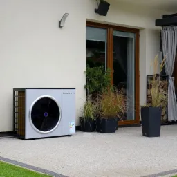 Szara pompa ciepła marki Rakentoo zainstalowana na zewnątrz budynku, obok roślin w donicach i stosu drewna pod zadaszeniem.