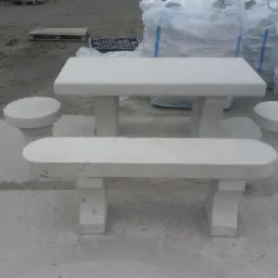 Komplet mebli ogrodowych z jasnego granitu: stół i dwa siedziska. W tle worki z kostką granitową i palety. Surowy, minimalistyczny design.