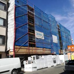 Sevo - Budynek w rusztowaniach, osłonięty niebieską siatką, z widocznym banerem reklamowym firmy Van Huele, obok zaparkowany biały samochód dostawczy i palety materiałów izolacyjnych.