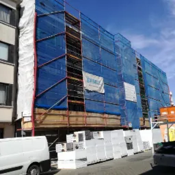 Budynek w rusztowaniach, osłonięty niebieską siatką, z widocznym banerem reklamowym firmy Van Huele, obok zaparkowany biały samochód dostawczy i palety materiałów izolacyjnych.