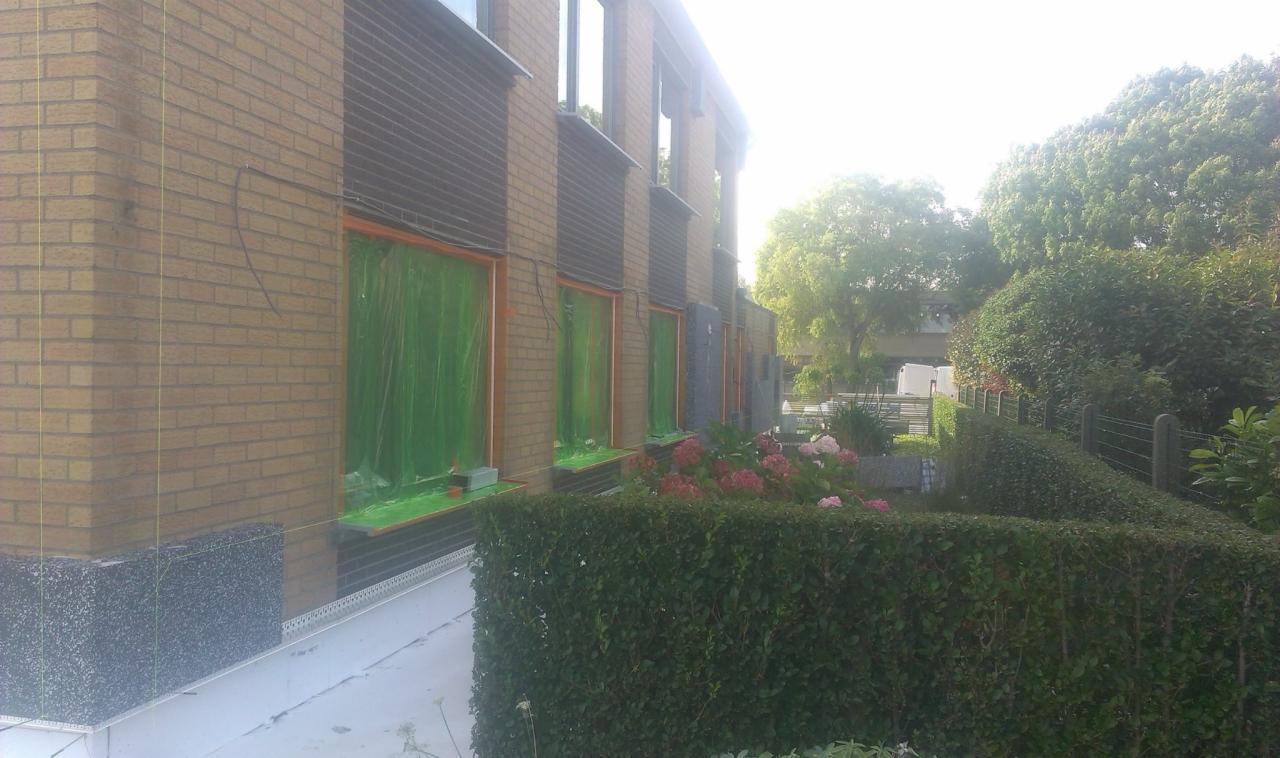 Budynek z cegły z oknami zabezpieczonymi folią ochronną, fragment elewacji z szarym ociepleniem, widoczne linki pionujące, krzewy i kwiaty w ogrodzie.