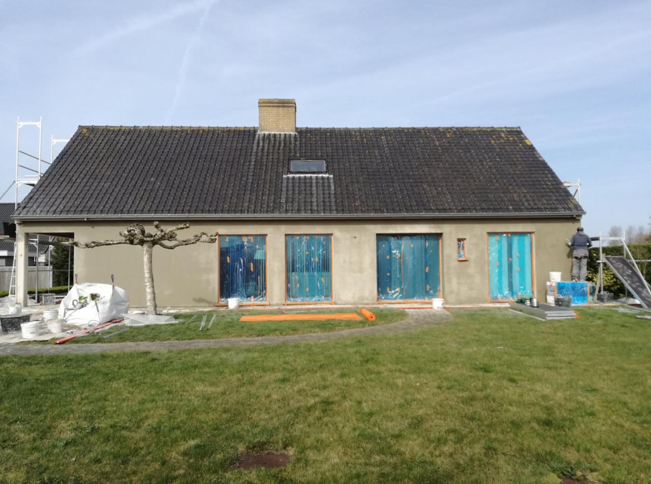 Dom jednorodzinny w trakcie tynkowania elewacji, okna zabezpieczone folią, widoczny pracownik na rusztowaniu i materiały budowlane na trawniku.