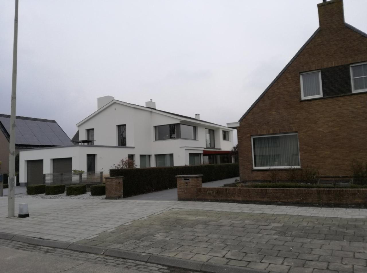 Nowoczesny dom z białą elewacją i dużymi oknami, widoczny z perspektywy ulicy, z domem z cegły w tle po prawej stronie.