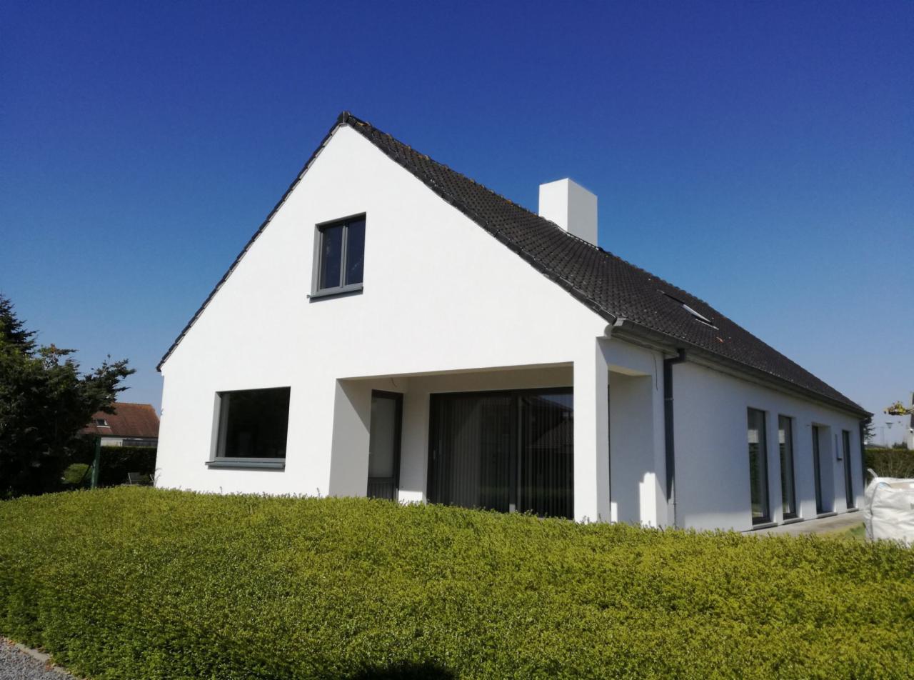 Nowoczesny dom jednorodzinny z białą elewacją i grafitowymi oknami, otoczony zielonym żywopłotem, widok w słoneczny dzień.