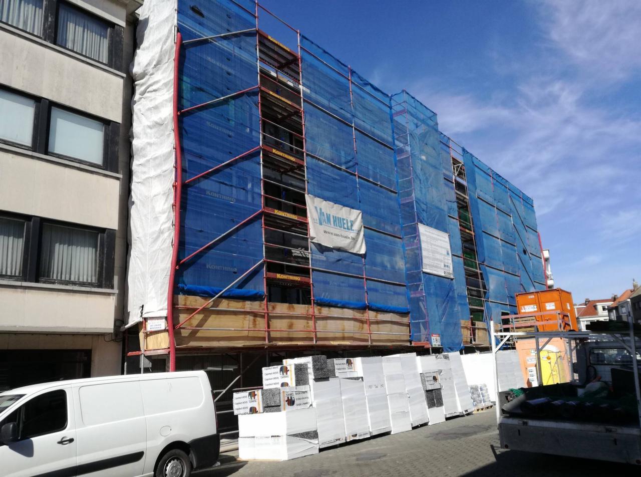 Budynek w rusztowaniach, osłonięty niebieską siatką, z widocznym banerem reklamowym firmy Van Huele, obok zaparkowany biały samochód dostawczy i palety materiałów izolacyjnych.