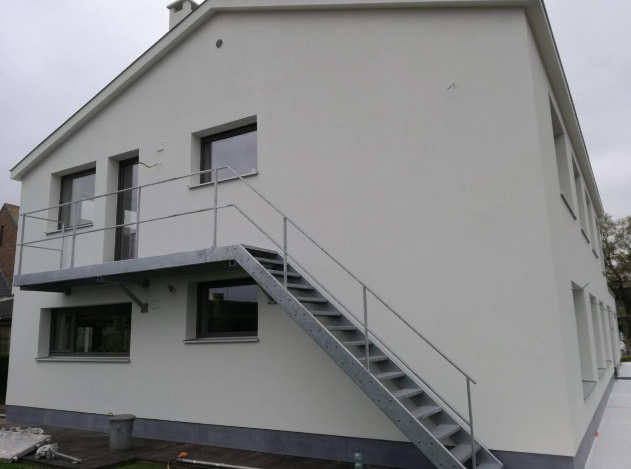 Jasna elewacja budynku z zewnętrznymi schodami metalowymi prowadzącymi na balkon, widoczne okna z ciemnymi ramami.