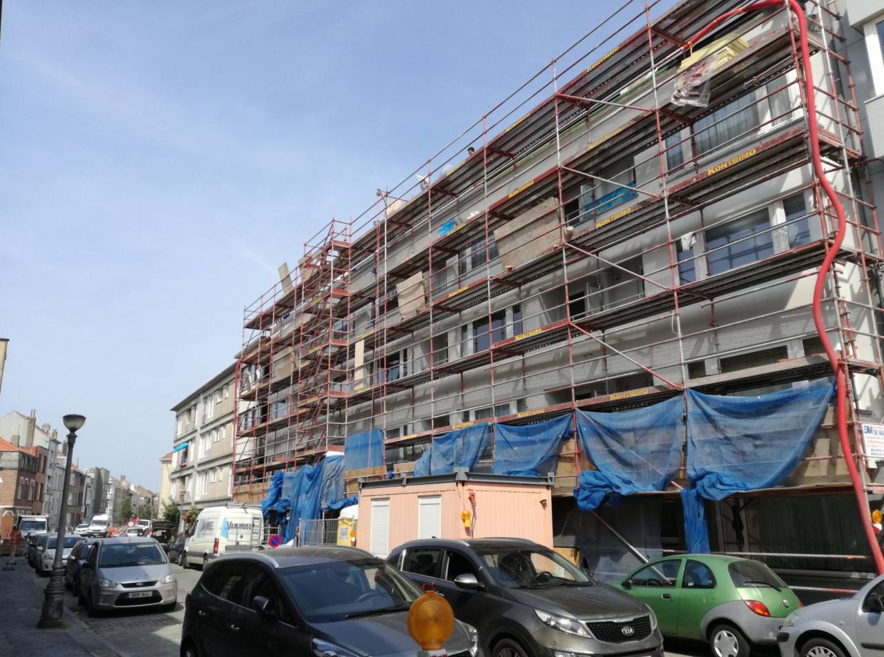 Wielopiętrowy budynek w trakcie prac elewacyjnych, otoczony rusztowaniami i zabezpieczony niebieską siatką, z widoczną czerwoną rurą doprowadzającą materiał, oraz zaparkowanymi samochodami na ulicy.