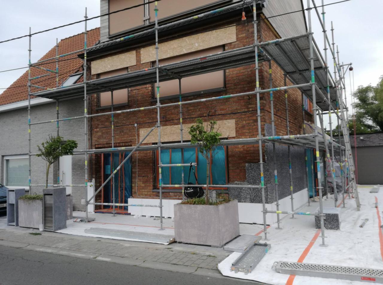 Dom z cegły w trakcie ocieplania, widoczne rusztowania, styropian grafitowy, zabezpieczone okna i drzwi folią, donice z drzewkami na pierwszym planie.