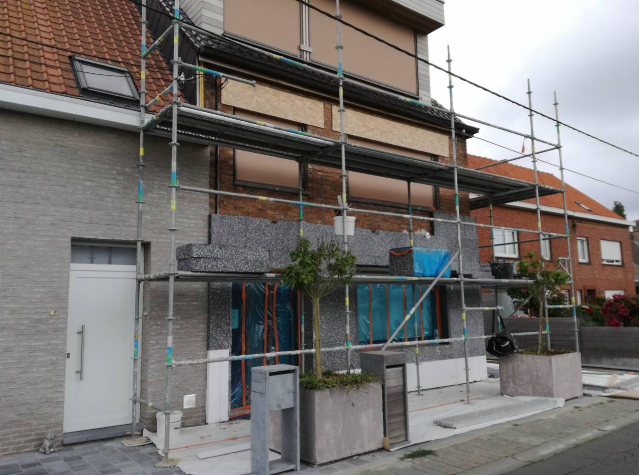 Dom w trakcie ocieplania styropianem grafitowym, widoczne rusztowania, zabezpieczone okna folią, skrzynki pocztowe i donice z roślinami przed budynkiem.
