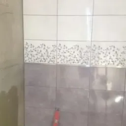 Układanie płytek w łazience: widoczne białe płytki z motywem roślinnym u góry oraz szare płytki poniżej, pionowy poziomnica przy ścianie, surowy beton na ścianie obok.