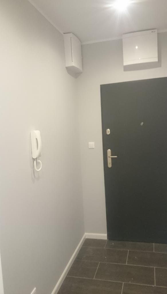 Szare wnętrze przedpokoju z zamontowanym klimatyzatorem na ścianie, obok drzwi wejściowych i domofonu.