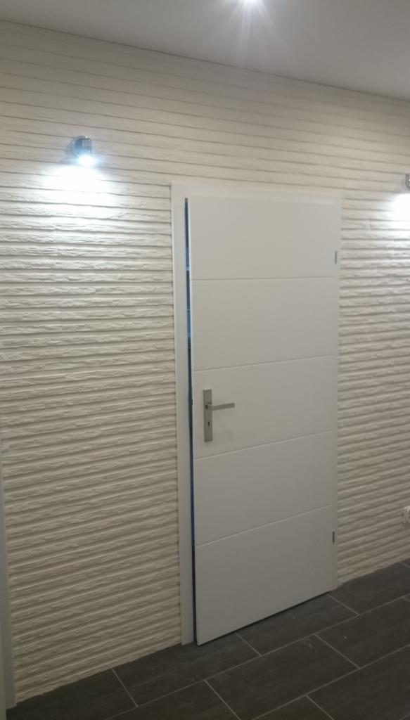 Białe, minimalistyczne drzwi wewnętrzne z poziomymi przetłoczeniami, zamontowane w ścianie wykończonej dekoracyjną, poziomą fakturą, oświetlone dwoma punktowymi lampami.