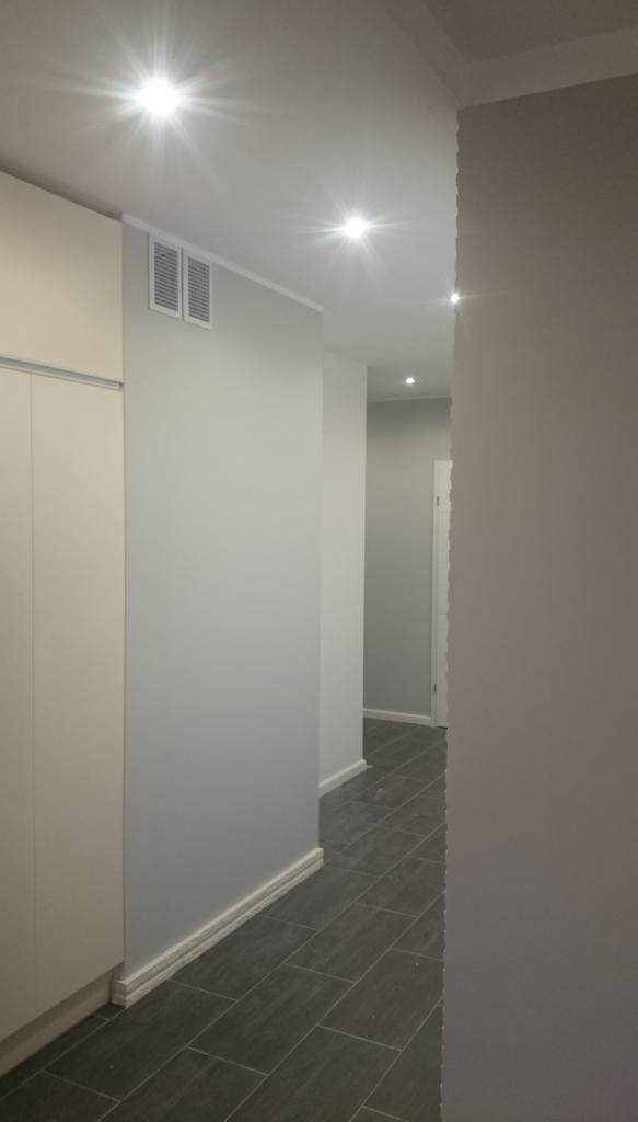 Korytarz w odcieniach szarości z ciemną podłogą z płytek imitujących drewno, oświetlony punktowymi lampami sufitowymi i wykończony białymi listwami przypodłogowymi.