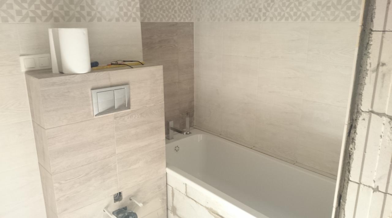 Biała wanna obudowana płytkami imitującymi drewno, z zamontowaną baterią i przyciskiem spłuczki; widoczne fragmenty surowej ściany i dekoracyjne wykończenie górnej części ściany.