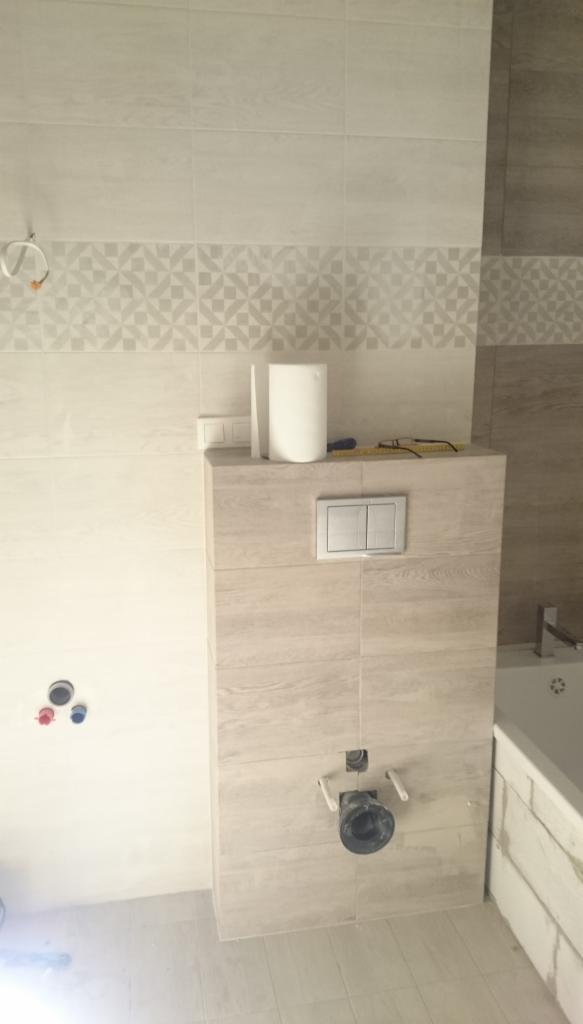 Ściana w łazience wyłożona jasnymi płytkami z dekoracyjnym pasem, zabudowa stelaża podtynkowego WC z przyciskiem spłukującym, widoczne elementy instalacji hydraulicznej i fragment wanny.