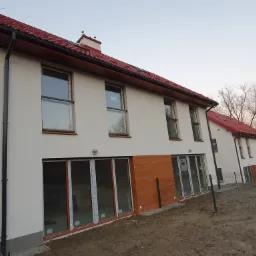 Nowoczesny, dwukondygnacyjny budynek z białymi ścianami i czerwoną dachówką w trakcie wykończenia, z widocznymi drewnianymi elementami elewacji i ogrodzeniem panelowym.