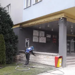 Pracownik w kombinezonie ochronnym czyści kamienną elewację budynku użyteczności publicznej za pomocą urządzenia wysokociśnieniowego, widoczny efekt czyszczenia i otoczenie budynku.