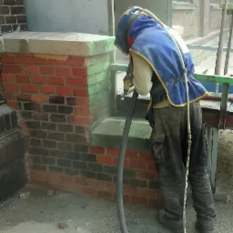 Pracownik w kombinezonie ochronnym czyści ceglany narożnik budynku za pomocą strumienia pod ciśnieniem, widoczne efekty czyszczenia na cegłach.