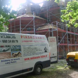 Renowacja ceglanej elewacji zabytkowego budynku sakralnego, widoczne rusztowania, samochód dostawczy z reklamą firmy oferującej usługi czyszczenia strumieniowo-ściernego i sodowania oraz żółty...