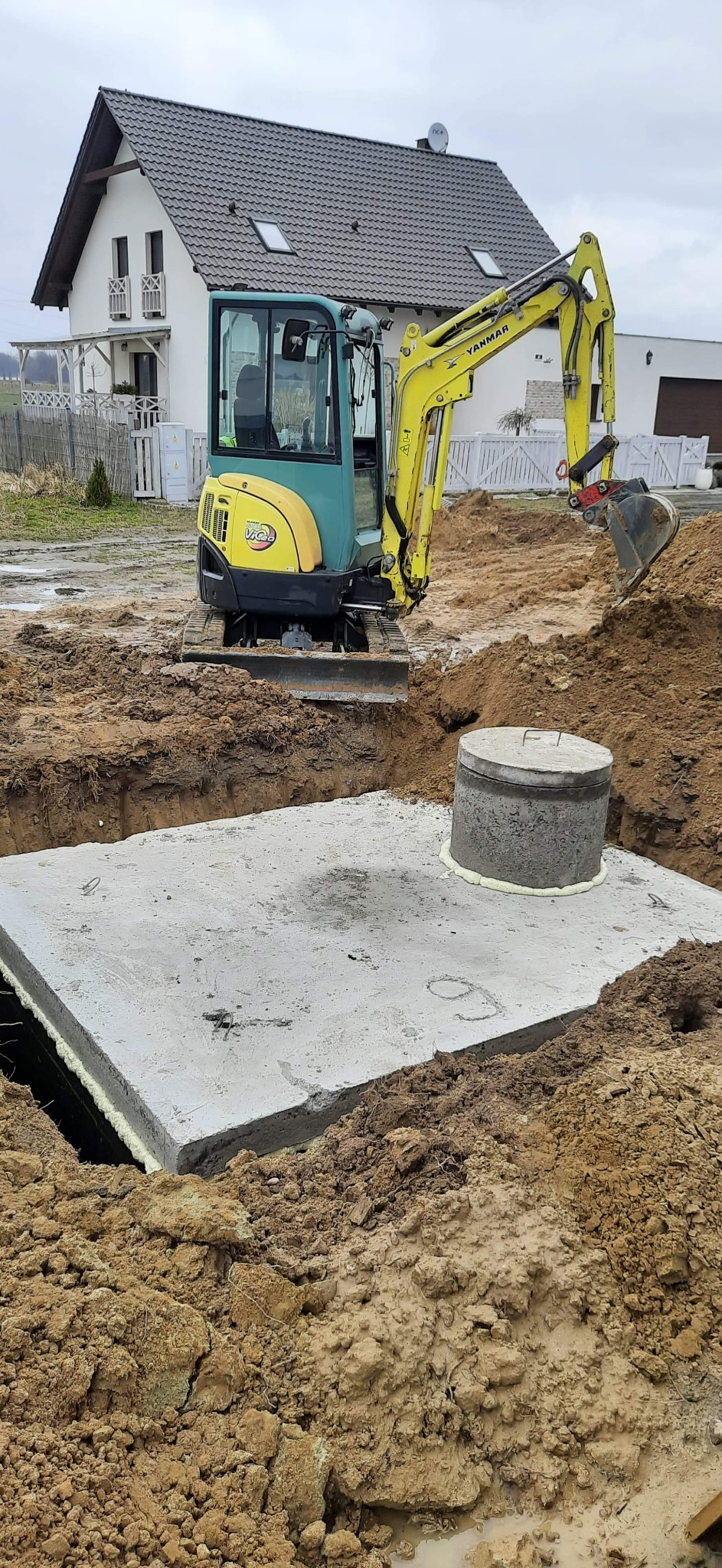 Żółto-zielona mini koparka Yanmar obok betonowego zbiornika z pokrywą na placu budowy domu jednorodzinnego, ziemia wokół wykopu zabezpieczona pianką montażową.
