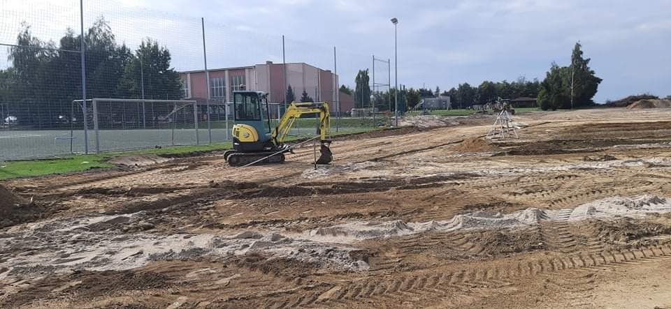 Żółto-szara mini koparka na placu budowy, przygotowanie terenu pod budowę boiska sportowego, widoczne ślady opon, budynek w tle.