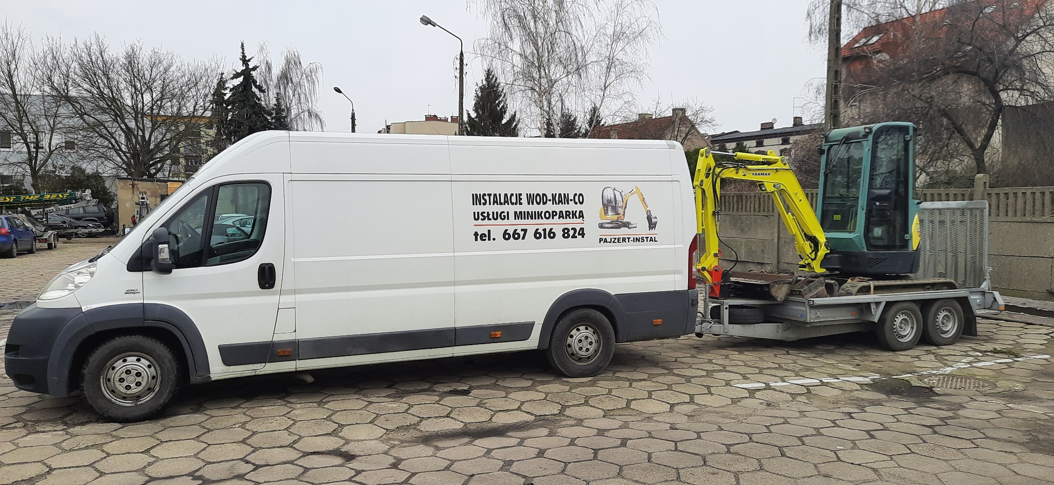 Biały samochód dostawczy z reklamą usług minikoparką, ciągnący przyczepę z żółto-zieloną minikoparką Yanmar, zaparkowany na parkingu z kostki brukowej.