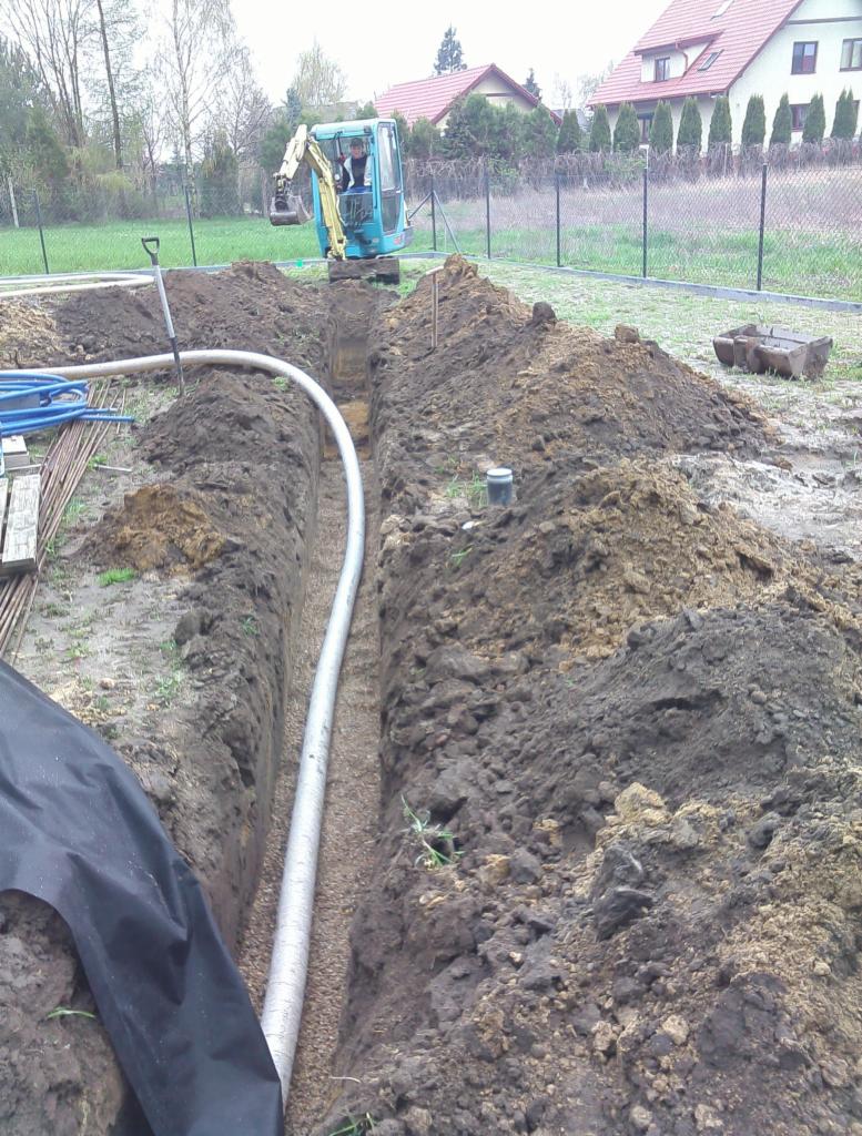 Wykopany rów z ułożoną rurą drenarską obsypaną żwirem, widoczna mała koparka i operator w kabinie, teren budowy w pobliżu domów jednorodzinnych.
