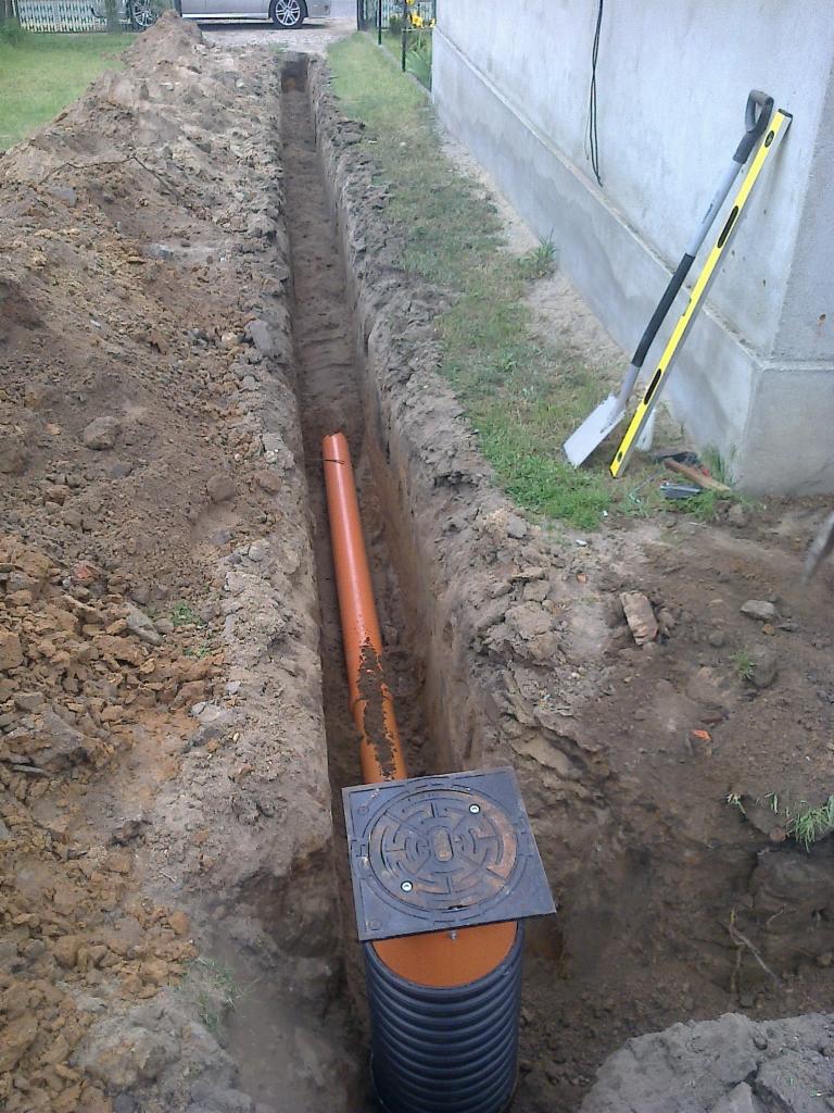 Wykop z rurą kanalizacyjną i studzienką rewizyjną, obok ściany budynku. Na ziemi leżą szpadel i poziomica. Widok z góry na wykop.