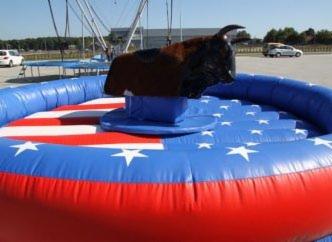 Symulator byka rodeo na dmuchanym podeście w kolorach flagi USA, widoczne elementy konstrukcji i pojazdy w tle.