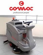 Szorowarka do posadzek marki Comac, model nieznany, na jasnym tle z widocznym oknem w tle. Maszyna w kolorze szarym z czerwonym logo producenta u góry.