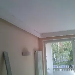 Ściana w pokoju z zamontowaną listwą dekoracyjną pod sufitem, widoczne ślady po szpachlowaniu. Okno balkonowe z widokiem na zieleń.