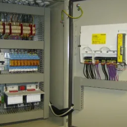 Szara szafa sterownicza z widocznymi elementami elektrycznymi, w tym przekaźnikami, bezpiecznikami i przewodami o różnych kolorach, zamontowana na ścianie.