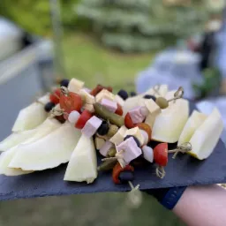Szpikulce z melonem, pomidorkami cherry, oliwkami, og&oacute;rkami konserwowymi i kostkami szynki podane na czarnym, kamiennym talerzu trzymanym w dłoni na tle zieleni ogrodowej i cateringu.