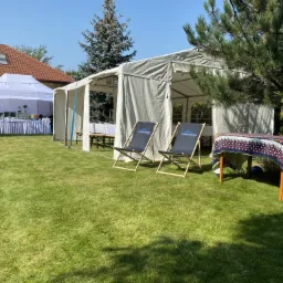 Ustawione na trawie dwa namioty: jeden biały z bufetem, drugi kremowy z ławkami wewnątrz. Dwa leżaki z logo firmy stoją przed namiotem. Stół z kolorowym obrusem i butelkami alkoholu z prawej strony...