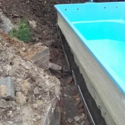 Montaż gotowego basenu w ziemi, widoczny wykop z deskowaniem, częściowo obsypany ziemią i gruzem, w tle mała koparka CAT 301.8C.