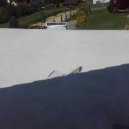 Świeżo wylany, gładki strop betonowy z widocznym cieniem barierki, w tle zaparkowany biały samochód dostawczy i zieleń ogrodu.