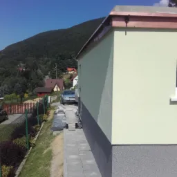 Świeżo otynkowana elewacja domu z dwukolorowym wykończeniem, widoczne elementy orynnowania z miedzi, w tle zalesione wzgórze i inne zabudowania.