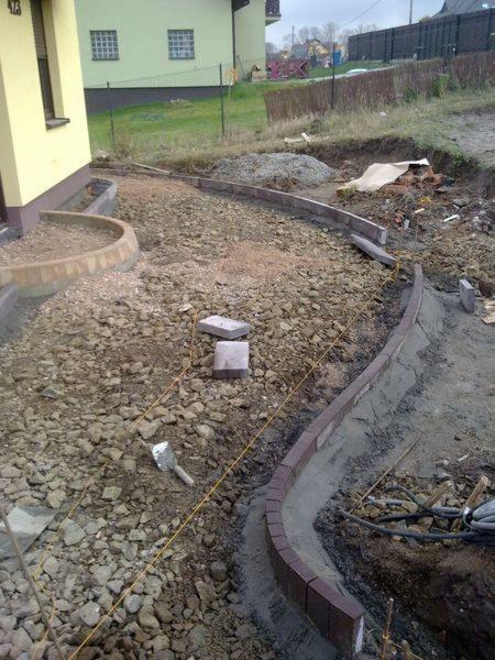 Przygotowanie podłoża pod ścieżkę z obrzeżami z kostki, widoczne kamienie, piasek, sznurek wyznaczający trasę, narzędzie murarskie i fragment budynku w tle.