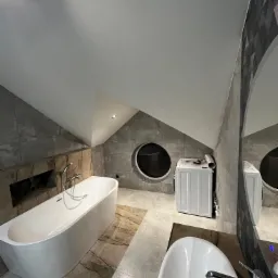 Nowoczesna łazienka na poddaszu z owalną wanną, umywalką nablatową i okrągłym oknem, wykończona szarą płytką imitującą kamień i beżowymi płytkami podłogowymi.