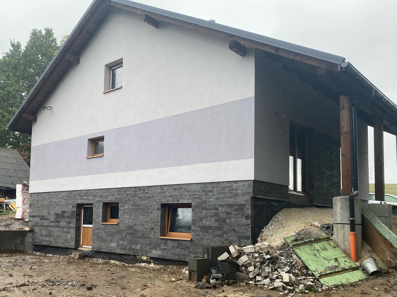 Dwukondygnacyjny dom z szarą elewacją podzieloną poziomym pasem, dolna część wykończona kamieniem elewacyjnym, widoczne okna z drewnianymi ramami oraz elementy rusztowania i pozostałości po pracach...