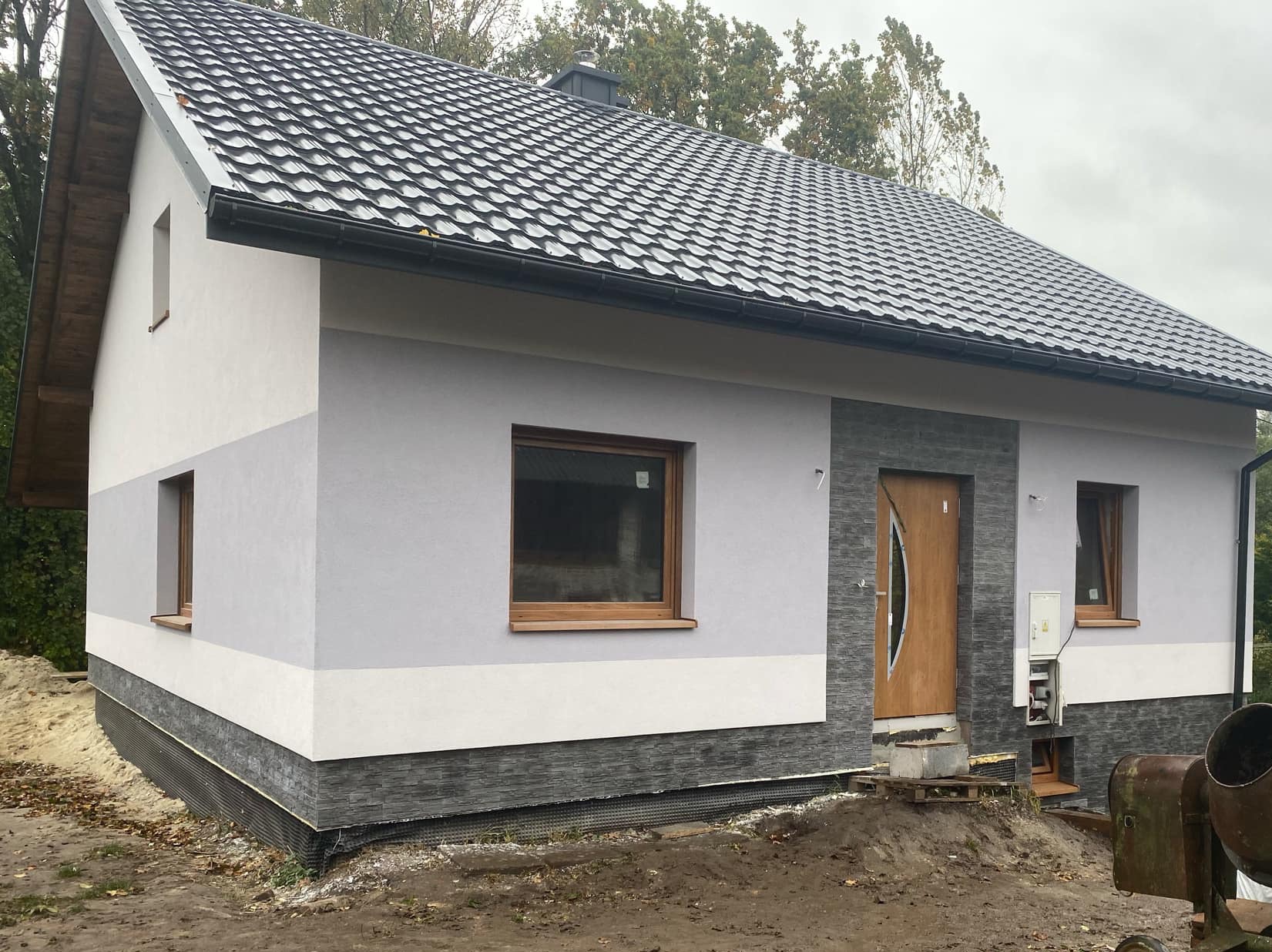 Nowo wybudowany dom jednorodzinny z szarą elewacją podzieloną na trzy pasy kolorystyczne, z ciemnoszarą podmurówką, drewnianymi oknami i drzwiami wejściowymi z numerem 7, widoczny mikser budowlany.