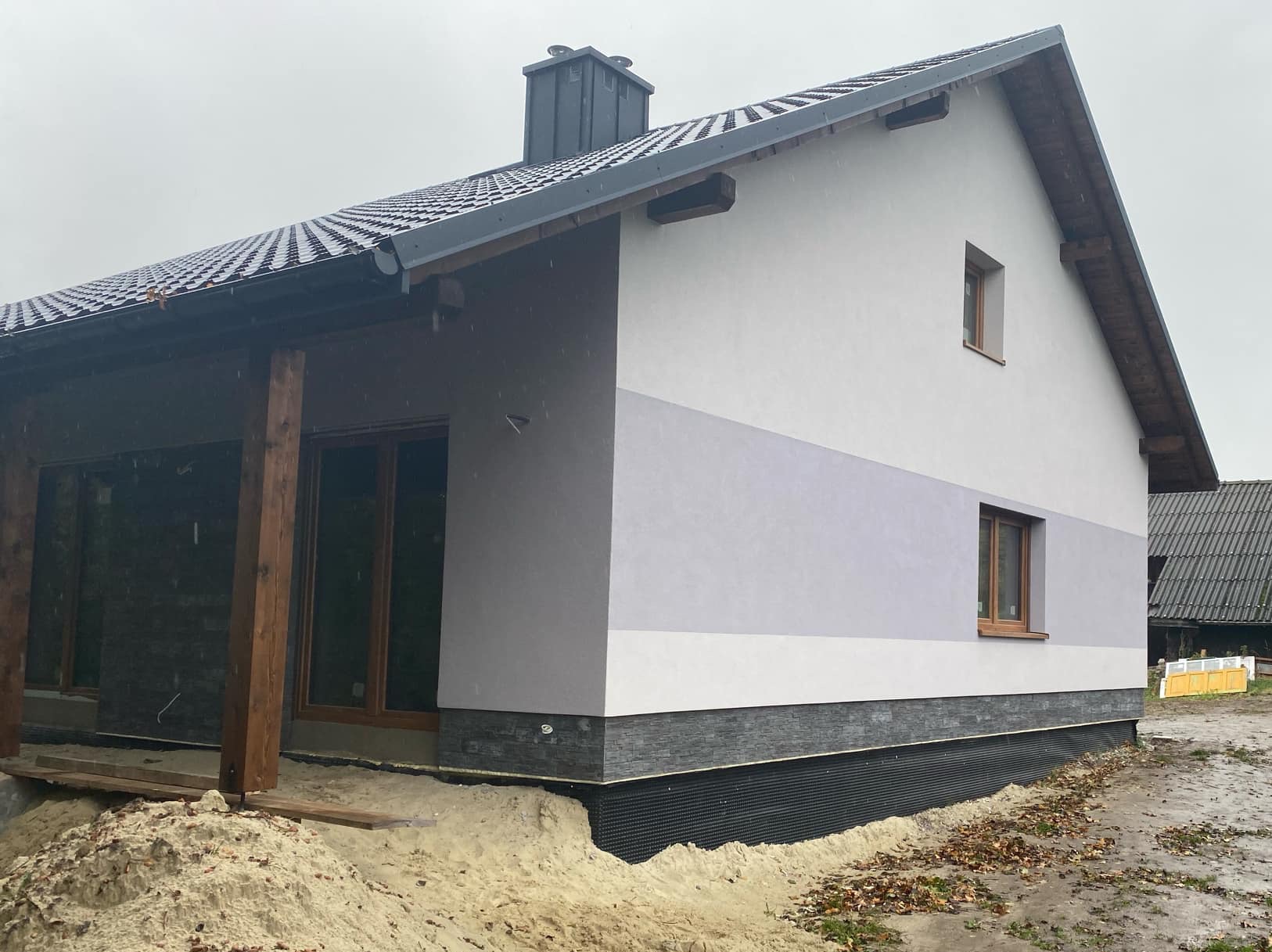 Nowoczesny dom jednorodzinny z elewacją w odcieniach szarości i bieli, wykończony kamieniem elewacyjnym u podstawy, widoczne drewniane elementy konstrukcyjne i okna z drewnianą stolarką, teren...