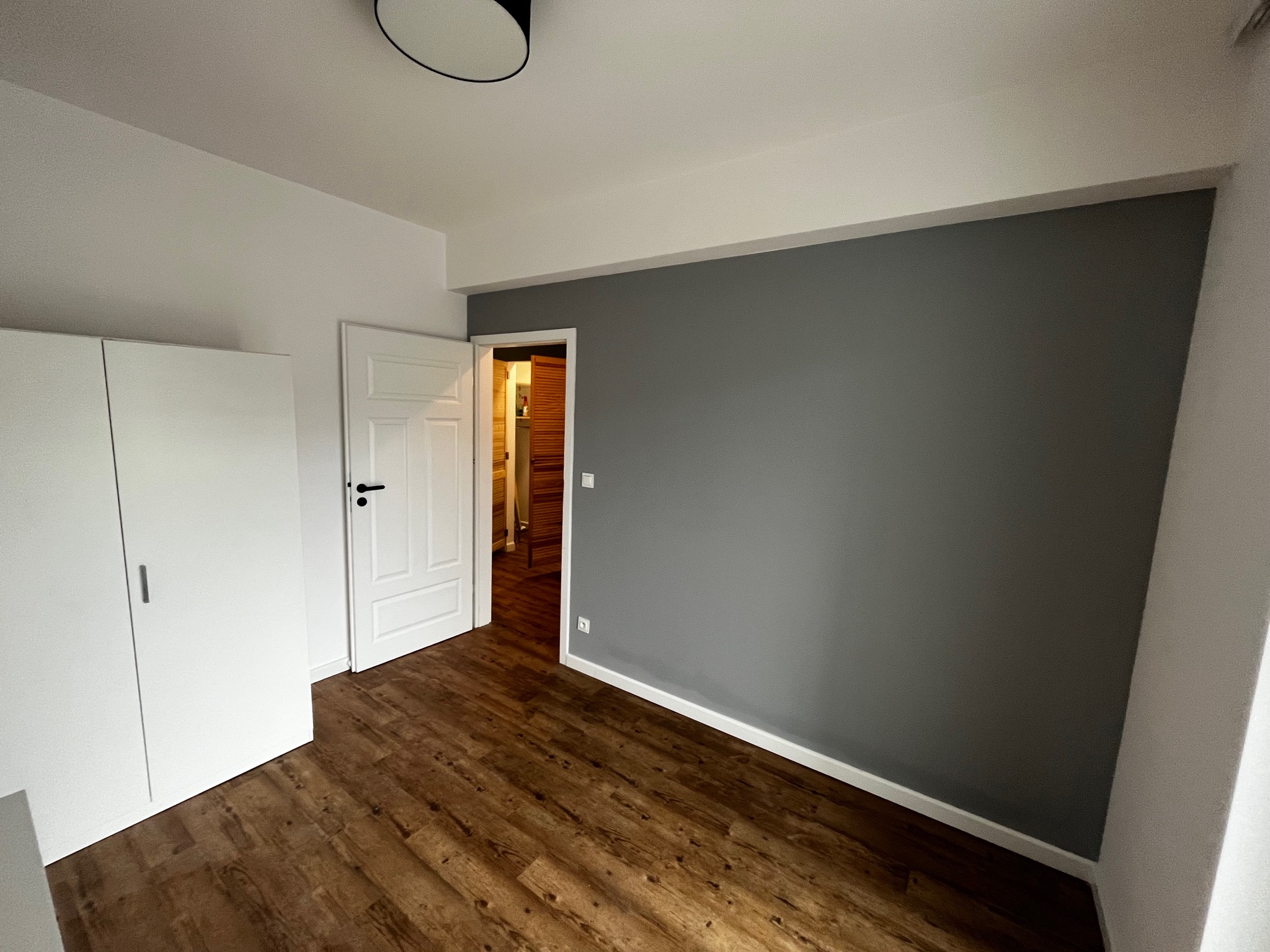Pusty pokój po remoncie: szara ściana, drewnopodobna podłoga, białe drzwi i szafa w minimalistycznym stylu.