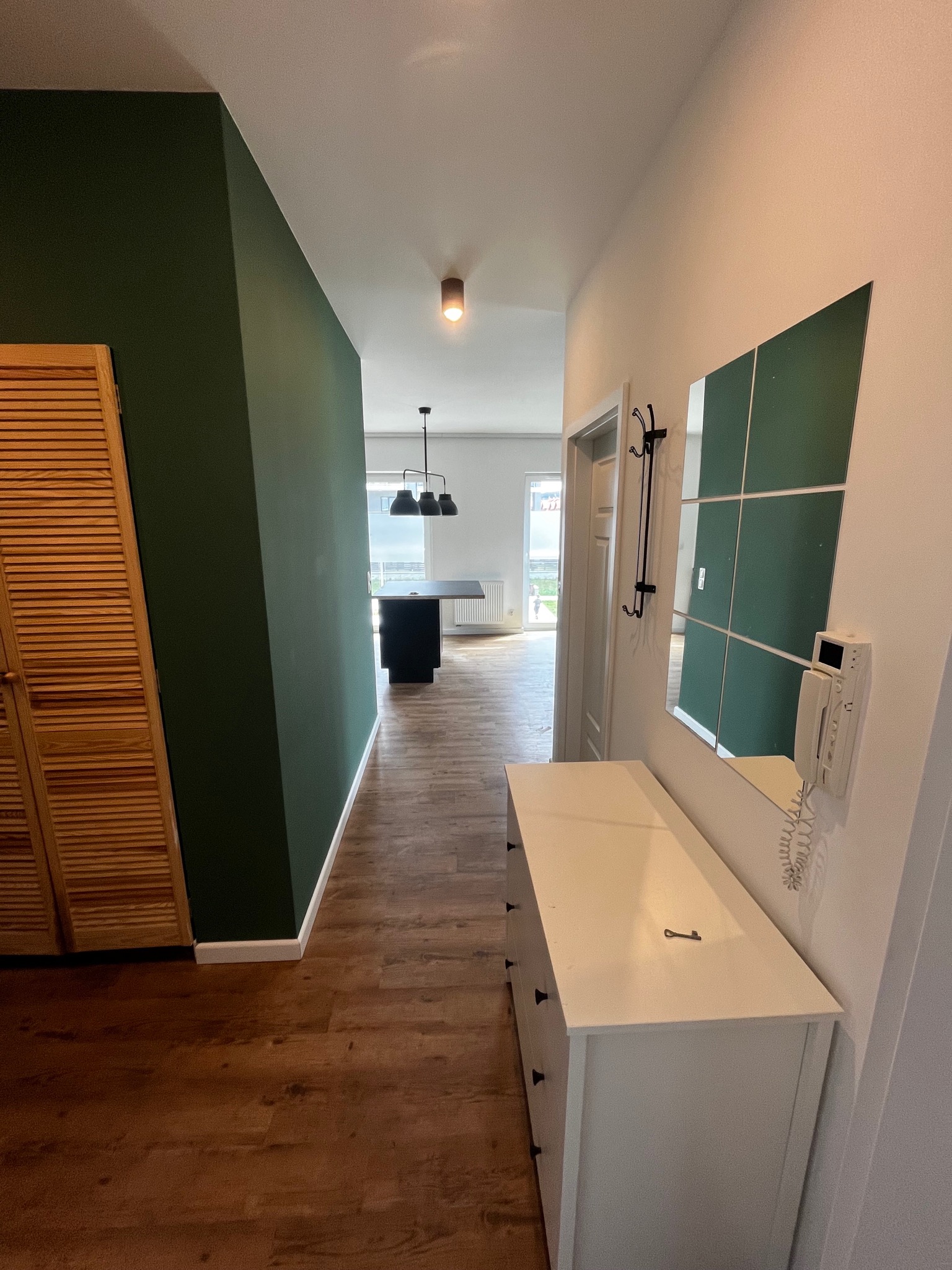 Nowoczesny przedpokój z ciemnozieloną ścianą, drewnianą szafą z żaluzjowymi drzwiami, komodą z lustrem w ramie z zielonymi kwadratami, drewnianą podłogą i widokiem na kuchnię z czarną wyspą i lampami.