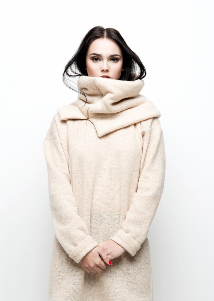 Brunetka w beżowej tunice z golfem zapinanym na zamek, na białym tle, z dłońmi splecionymi na wysokości bioder, prezentuje jesienną kolekcję.