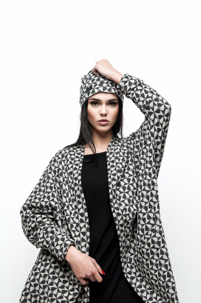 Kobieta w czarnej sukience i wzorzystym kardiganie oraz czapce. Ujęcie od dołu, białe tło, profesjonalna sesja zdjęciowa. Wyrazista stylizacja na jesień.