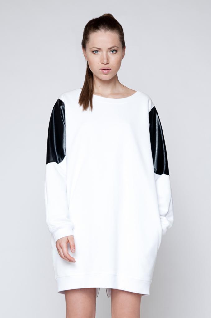 Kobieta prezentuje białą sukienkę dresową z długim rękawem i czarnymi, błyszczącymi wstawkami na ramionach na jasnym tle.