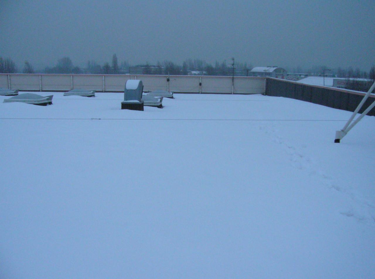 Zasypany śniegiem płaski dach budynku z elementami wentylacyjnymi i śladami stóp. Widok na zabudowania w tle, pochmurne niebo.
